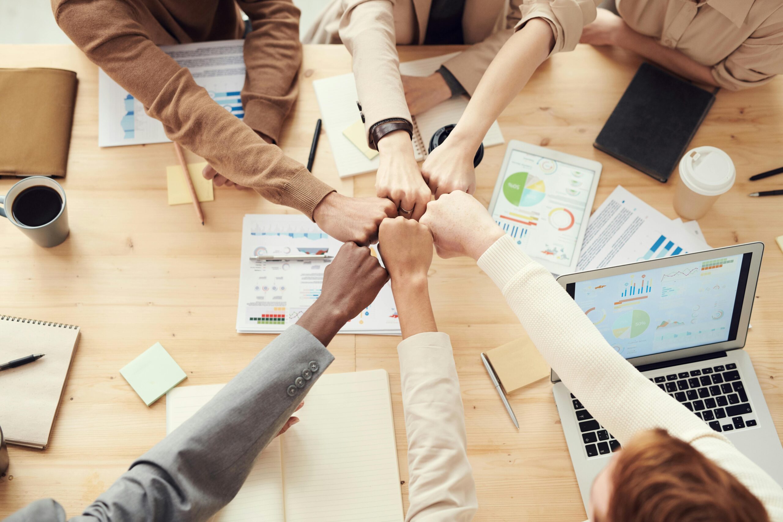 Top view of a diverse team fist bumping over a meeting table with laptops and paperwork, highlighting unity, collaboration, and shared goals.